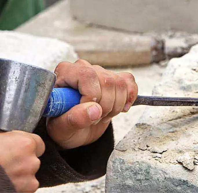 La main d'une personne tenant un ciseau de maçon et un marteau, en train de travailler sur un bloc de pierre, en sculptant ou en façonnant la pierre.
